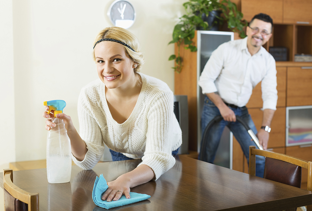Positive young spouses dusting and hoovering at domestic interior